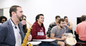 Foto de uma sala de aula. Mais a esquerda, o professor está sentado e tocando um saxofone, ao lado outros alunos estão sentados nas carteiras e olham para frente. Um deles segura um pandeiro e, outro, ao fundo, uma flauta.
