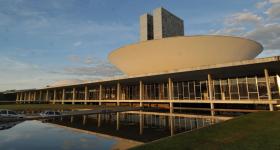 Foto do Congresso Nacional, mostrando em primeiro plano o espelho d'água, as janelas envidraçadas e a cúpula convexa da Câmara dos Deputados. Ao fundo, as torres centrais. 