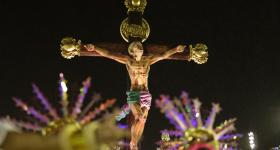  Foto de uma das alegorias apresentadas pela Estação Primeira de Mangueira durante o desfile das escolas de samba do Rio de Janeiro, no Carnaval de 2020. O carro alegórico faz uma releitura da história de Jesus Cristo, como um jovem negro baleado. Na imagem, o jovem está crucificado e olha para o céu. Acima, na cruz, lê-se a palavra "negro". Abaixo, aparecem membros da escola fantasiados, em primeiro plano mas desfocados. 