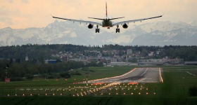 Foto de avião pousando em uma pista de aeroporto. Observa-se a parte traseira do avião, bo centro da imagem, que é branco e possui detalhes em laranja. A pista de pouso está imulinada com luzes amarelas e laranjas e ao redor há terrenod com grama e árvores baixas. Em segundo plano, há uma uma cidade, onde nota-se prédios e árvores e, ao fundo, diversas montanhas.
