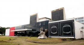 Foto da fachada do Espaço das Artes. Prédio térreo e retangular. à esquerda, uma pintura de formas geométricas, dois quadrados e circulos na cor branca em fundo preto, ao centro, o prédio possui vidro preto e na ponta, uma parede na cor vermelha. Uma obra de arte em metal está a frente do prédio.