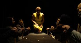 Foto de cena do espetáculo oresteia.br. Uma mulher está semi ajoelhada sobre uma mesa cercada por homens vestindo terno e usando máscaras. A mulher usa um vestido amarelo, tem pele clara e cabelos cacheados e curtos. A luz principal incide do teto, criando sombras e contrastes intensos.