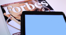 Foto de uma revista e um tablet sobre uma mesa. Em primeiro plano, o tablet, com a tela em branco. Sob o tablet, uma edição da revista Forbes. 