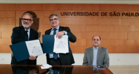 Foto do professor Paulo Nassar, um homem branco de cabelos e barba branca e óculos, e do reitor Carlos Gilberto Carlotti Jr, um homem branco de óculos e cabelos grisalhos, posando com exemplares do convênio entre ECA e Aberje. Ambos estão de pé atrás de uma mesa de madeira. Sentado na ponta da mesa, o professor Eugênio Bucci, um homem branco e calvo de cabelos escuros. Todos usam paletó e sorriem para a câmera.