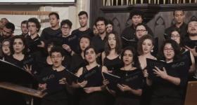 Foto de parte do Coral da ECA-USP, onde estão agrupadas várias fileiras de homens e mulheres cantando com uma pasta nas mãos
