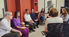 Foto de pessoas reunidas em um semi-circulo, parte delas sentada em um banco de madeira e encostados em um parede vermelha, outra parte em cadeiras de escritório. São três homens e quatro mulheres.