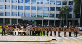  Foto de estudantes e professores em frente ao prédio da reitoria da USP. Vários estudantes, brancos e negros, estão alinhados lado a lado e seguram um cartaz bege em que está escrito "Cadê os docentes pretos e indígenas?”. Atrás dos estudantes, há um prédio espelhado