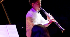 Foto de um homem tocando clarinete. Ele é branco, tem cabelos curtos e escuros e veste uma camisa branca. O fundo da imagem é totalmente preto, e o jovem é iluminado por luzes branca e roxa. Na porção inferior da foto, aparecem a cabeça de uma mulher que está de costas para a câmera e uma partitura apoiada no piano que ela toca.