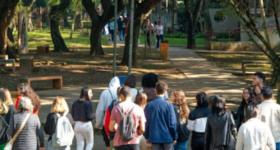  Foto tirada durante a visita monitorada à ECA. Na imagem, um grupo de estudantes passa pela parte externa da unidade, onde há várias árvores e bancos de concreto. Na foto, conta-se 19 pessoas andando de costas para a câmera.