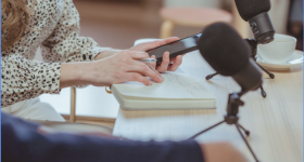 Foto de um trabalho jornalístico. Na imagem, aparecem as mãos de uma jornalista, que segura uma caneta e um celular, enquanto verifica anotações em um caderno. Na mesa em que está apoiada, há dois microfones e um conjunto de xícara e pires brancos. A jornalista está em segundo plano, e usa uma blusa de manga comprida branca com detalhes pretos. Ela é loira. Em primeiro plano, desfocado, está o braço de outra pessoa.