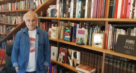 Foto de um homem posicionado em frente a uma estante de livros. Ele é branco, tem cabelos curtos e grisalhos e olhos escuros. Ele veste uma camiseta branca estampada e uma camisa jeans por cima. A estante é grande e ocupa toda a parede e, consequentemente, todo o fundo da imagem.