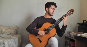 Foto de um rapaz tocando violão clássico e olhando para o instrumento. Ele é branco, tem cabelos curtos e escuros e veste uma blusa de manga longa preta e calça cinza. O jovem está em um quarto, sentado entre uma cama e uma escrivaninha. A parede ao fundo é cinza.
