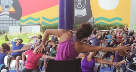 Foto de atividade no Sesc Pinheiros. Grupo de mulheres praticam atividade física. A professora está sentada em uma cadeira, de costas. Ela tem cabelos castanhos, ligeiramente ondulados, presos no topo da cabeça. Ela veste uma blusa roxa regata. à frente dela, está um grupo de mulheres sentadas em cadeiras, uma ao lado da outra. Elas repetem os movimentos da professora: um braço se estica para o lado, torcendo o tronco para o mesmo lado. o outro braço se mantém dobrado. Ao fundo, um painel artístico.