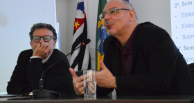 Foto de dois homens sentados em uma mesa. À esquerda, um homem de cabelos levemente grisalhos, usando óculos com haste escura, um terno preto e uma camisa branca, está com o queixo apoiado em uma das mãos. À direita, um homem calvo, de barba rala, usando óculos, um paletó preto e uma camisa escura fala ao microfone e gesticula com as mãos. Ao fundo, uma parede branca e as bandeiras do estado de São Paulo, do Brasil e da USP.