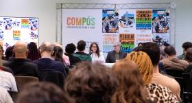 Foto de um evento acadêmico. Várias pessoas estão sentadas em fileiras enquanto olham para uma mesa à frente. Na mesa, uma mulher e um homem estão sentados de frente para o público. A mulher tem pele branca, usa óculos e veste roupas brancas, ela segura uma folha de papel e fala ao microfone. O homem, de pele branca, cabelos grisalhos, óculos e traje social, olha para a mulher ao seu lado. Um painel com a arte e identidade do evento e seu nome, Compós, está posicionado atrás deles. 