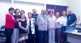 Foto de nove mulheres e três homens reunidos posando para uma foto de grupo. São todas pessoas idosas. Atrás das pessoas há uma porta dupla fechada, uma parede e é possível ver uma carteira de sala de aula. Também é possível ver uma mesa de professor com algumas vasilhas e sobras de comida. Aparentemente se trata de uma confraternização.