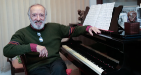 Foto de um homem branco, calvo, de cabelos e barba brancos. Ele usa um suéter verde escuro com punhos vermelhos e calça preta. Seus óculos de grau estão apoiados na gola do suéter. Ele sorri e posa ao lado de um piano de madeira escura, sobre o qual estende o braço esquerdo. Sobre a tampa do piano há uma partitura aberta..Ao fundo, uma persiana bege.