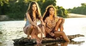 Foto de duas mulheres sobre um tronco de árvore em um rio. A da esquerda é branca, tem cabelos castanhos compridos, usa uma regata cinza e saia rosa e está agachada. A da direita está sentada, com uma perna apoiando no tronco e a outra mergulhada no rio. Seu braço esquerdo se apoia no joelho esquerdo. Ela tem a pele levemente bronzeada, cabelos castanhos escuros compridos, usa camiseta de manga curta em tons de marrom claro e saia marrom escuro. Ao fundo, a margem do rio e árvores.