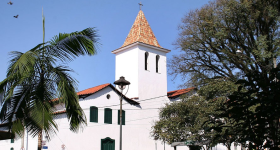 Foto da fachada de uma igreja em estilo colonial. As paredes são brancas, enquanto portas e janelas são da cor azul-marinho. Ao centro, a torre, que possui duas janelas estreitas e um telhado em forma de pirâmide com uma cruz no topo. A base da torre se liga, pelas laterais, a duas casas. Em primeiro plano, um poste de luz e as folhagens de uma árvore e de uma palmeira. Ao fundo, o céu azul, sem nuvens. 