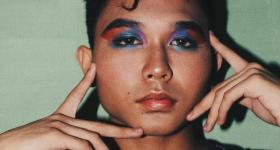 Foto de um rapaz com uma maquiagem azul e rosa nos olhos. Ele tem a pele bronzeada, cabelos lisos curtos e escuros, com uma pequena mecha na testa. Ele toca delicadamente o rosto com os dedos indicadores e médios  das duas mãos. Ele está com uma roupa escura com reflexos brilhantes da cor vinho