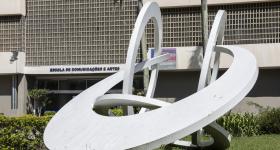 Foto de parte da fachada do prédio da ECA. Sobre um gramado verde, está uma escultura branca formada por quatro arcos que se entrelaçam entre si: um à frente, dois paralelos um ao outro e um quarto arco, mais próximo ao chão. Atrás da escultura, parte da fachado do prédio. A fachada tem elementos vazados, do tipo cobogó,  na cor bege. O nome da escola está afixado na parede, com as letras na cor azul escuro.