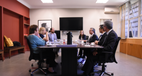 Foto de uma mesa de reuniões, com pessoas sentadas em cadeiras em volta dela. Ao fundo, um monitor de tv grande. Sobre a mesa, copos, xicaras e uma garrafa de café.