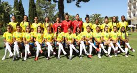 Foto oficial da Seleção Brasileira Feminina de 2019. 25 mulheres posam para uma foto, sendo 12 em pé e, à frente delas, 13 sentadas em um banco com as pernas cruzadas. Três das mulheres, que estão em pé e no centro da fotografia, usam um uniformes vermelhos, as demais usam uniformes amarelo e azul e meião branco. Elas estão em um gramado, a maioria sorri para a câmera e há um prédio branco de quatro andares no canto direito da foto. Árvores de diferentes formatos formam o segundo plano da imagem. 