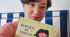 Foto da escritora Janaína Tokitaka, uma mulher de traços asiáticos, olhos castanhos, cabelos grisalhos, curtos e ondulados. Ela olha em direção à câmera e, com uma das mãos, segura seu livro Almoço de Família, cuja capa tem a ilustração de uma menina com cabelos cacheados e escuros, vestindo  blusa cor de rosa, em frente a uma mesa com pratos de comida. Ao lado da menina, na ilustração, há um gato.  Atrás de Janaína há um estofado com listras azuis e brancas e uma parede branca.