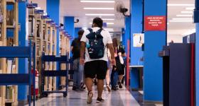 Um homem negro, vestindo uma camiseta branca e uma bermuda preta, caminha pelo corredor da biblioteca da ECA. Ele está de costas e carrega uma mochila. Do lado esquerdo, estantes de livros e, do lado direito, colunas, pintadas na cor azul. Outras pessoas caminham na sua frente