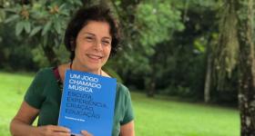Foto de uma mulher branca de cabelos curtos e escuros. Ela segura um livro com capa azul de título Um Jogo Chamado Música: escuta, experiência, criação, educação enquanto sorri e olha para o lado direito da imagem. Ao fundo, um gramado e folhagens de árvores. 