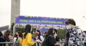 Foto de jovens andando em frente ao portal de entrada da feira. O banner sobre o portal é azul escuro, tem desenhos geométricos em tons de amarelo e rosa e nele está escrito “Feira USP e as Profissões 2022” em verde. Há diversos estudantes passando, eles usam máscara e a maioria está agasalhada. Ao fundo, aparece parte da Torre do Relógio e algumas árvores. O céu está nublado.