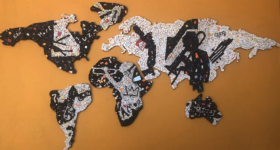 Foto da obra artística The World We Live In. Trata-se de um mapa mundi estilizado, com continentes formados por um mosaico no qual predominam peças brancas e pretas, pontuadas por peças em tons de laranja, amarelo, azul e vermelho. O contraste entre claro e escuro forma silhuetas de pessoas tocando trompete, saxofone e outros instrumentos. Há também desenhos de coroas, balanços e outros objetos, e palavras como “hope” e “amor”. O fundo, que representa os oceanos, é liso e de cor caramelo.