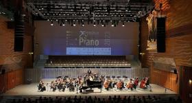 Foto do palco do 25o Concurso Internacional de Piano Santa Cecília. Na imagem, uma orquestra se apresenta para um público em uma sala de concerto. Ao centro do palco, um piano grande e preto. Ao fundo, no telão, aparece o cartaz de divulgação do concurso. A iluminação é direcionada ao palco e as paredes do ambiente têm tons de marrom e laranja. 