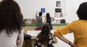 Foto de estudantes em um ateliê de arte. Ao fundo dois jovens, um homem e uma mulher, estão de pé, observando trabalhos de arte que estão afixados na parede e sobre uma bancada. Atrás deles, outras quatro jovens, também de costas, os observam. Elas estão sentadas em cadeiras e mesas do espaço.