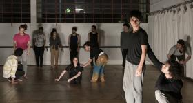 Foto. Em uma sala ampla com piso de madeira, paredes brancas, vitrô ao fundo e uma cortina branca no canto direito, um grupo de pessoas participa de uma aula de dança e expressão corporal. À frente, estão dois homens, jovens, brancos, com cabelos castanhos e ondulados; um está de pé e olha para baixo com expressão séria, o outro está agachado, erguendo um dos braços e tocando nas costas do primeiro. Eles se vestem de forma semelhante: calça cinza e blusa preta, descalços