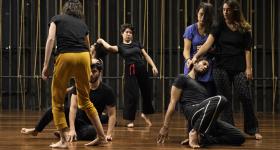 foto de sete jovens movendo-se em um palco. Eles são, na maioria, brancos,estão descalços e usam calças e camisetas de tecidos confortáveis e modelagem larga. A maioria das roupas é da cor preta, destacando-se uma calça amarela, uma camiseta azul e uma fita vermelha. Dois jovens estão semi ajoelhados, com as mãos tocando o piso, enquanto outros tocam suas cabeças com os braços esticados.  O piso é de madeira e no fundo escuro estão as cordas verticais que fazem a mecânica cênica.