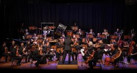 Foto de uma apresentação da Orquestra Parassinfônica de São Paulo. Pessoas, a maioria sentadas, vestidas de preto, tocam instrumentos como violoncelo, viola e violino em um palco de madeira. No centro, de costas para a câmera, um regente, com terno preto, movimenta os braços. A iluminação se concentra nos musicistas, ao redor deles há pouca luz. No fundo, uma cortina azul escuro. 