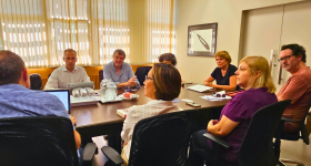 Foto de um grupo de pessoas sentado ao redor de uma mesa retangular. Na frente, de costas ou de lado para a foto, estão os docentes da ECA: dois homens e duas mulheres, sentados em cadeiras de escritório, na cor preta. Ao fundo, e de frente para a foto, estão três docentes da faculdade francesa. Na cabeceira da mesa, está sentada a diretora da ECA. Em cima da mesa de madeira, na cor marrom escuro, estão diversos objetos, jarras de água, copos de vidro, computadores, papeis, livros, jarra de café.