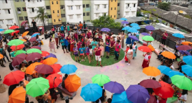 Foto de diversas pessoas reunidas na área externa de um conjunto habitacional. Elas formam uma roda, com diversas pessoas usando roupas coloridas e portando guarda-chuvas nas cores azul, roxo, verde, vermelho, vinho e laranja. No centro da roda, um jovem branco de cabelos escuros, camiseta vermelha e bermuda branca fala e gesticula. Ao lado dele há crianças e jovens, muitos deles negros. Ao fundo, parte da fachada de alguns prédios. 