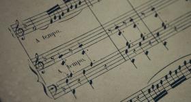 Foto de uma folha de partitura impressa. O papel é levemente amarelado, com algumas manchas. Sobre ele, parte das linhas da partitura, divididas em blocos de cinco linhas cada, que se alternam, no sentido horizontal, com espaços em branco. Sobre as linhas há diferentes notações musicais e em dois dos espaços em branco aparecem os dizeres “a tempo”. Todos os elementos impressos são da cor preta. 
