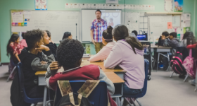 Fotografia de uma sala de aula com parede verde, piso cinza, quadro branco e cartazes. Estudantes  estão sentados em grupos em carteiras dispostas na sala com a atenção voltada ao professor, um homem branco que veste camisa xadrez e está à frente do quadro branco. 