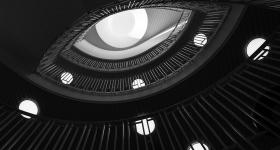 Foto preto e branco com a visão de baixo para cima do interior de um edifício. Há três andares com gradis em formato de elipse, que diminuem de tamanho com a visão em perspectiva de altura. Cada andar da construção tem várias lâmpadas brancas. Há uma esfera branca no topo do teto, onde o andar superior é mais claro e a cor predominante do entorno da foto é preta. Esse detalhe, somado ao formato dos gradis, lembra a imagem de um olho. 