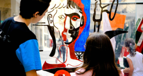 Uma criança de cabelos compridos e blusa Rosa e uma joem de cabelos curtos e blusa preta estão em pé, de costas, observando uma obra de arte, o desenho do busto de um homem, feito em materiais diversos, nas cores preto, vermelho azul e transparência. Ao fundo, prédios e a calçada mostram que elas estão em um espaço aberto da cidade de São Paulo