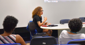 Foto de jovem negra, com cabelos longos, crespos e castanho-claros, usando óculos de haste escura e roupa preta. Ela está sentada em uma cadeira, de lado, e gesticula com uma das mãos enquanto se apresenta para pessoas que estão à sua frente. Duas outras pessoas, sentadas de costas, observam a apresentação.