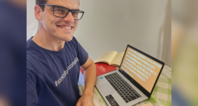 Foto de um jovem de pele branca, cabelos castanhos escuros e óculos de armação, vestindo uma camiseta azul escura. Ele está sentado e com o antebraço apoiado em uma mesa. à sua frente e em cima da mesa está um notebook e alguns livros. A parede ao fundo é branca