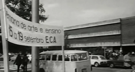 Captura de vídeo. A imagem é em preto e branco e mostra em primeiro plano, uma faixa de tecido com os dizeres Semana de Arte e Ensino. 15 a 19 de setembro. ECA. Atrás da faixa estão alguns carros e a fachada do Prédio Central da ECA, uma construção com três pavimentos marcados por três lajes horizontais intercaladas por elementos vazados à frente das janelas.