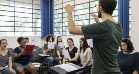 Foto de um jovem branco, de cabelos curtos e castanhos e barba da mesma cor, está de pé, com os braços ao alto. à sua frente uma partitura e mais ao fundo, um grupo de jovens estão sentados, com folhas nas mãos, cantando. Ao fundo, as janelas da sala de aula.