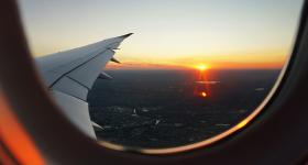 Foto da asa de um avião tirada pelo lado de dentro do avião. Na foto aparece uma extensa faixa de solo e um céu com tons de laranja e azul com o Sol no horizonte