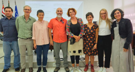 Foto de sete pessoas de pé em uma auditório, lado a lado, posando. Ao fundo, uma parede branca, com uma projeção, e as bandeiras de São Paulo, Do brasil e da USP, ao fundo, do lado esquerdo.