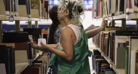 Foto de duas mulheres entre duas prateleiras de livros de uma biblioteca. Em primeiro plano, virada para a esquerda é branca, tem cabelo ondulado e castanho com mechas loiras, usa brinco de argolas, uma regata branca e uma jaqueta verde. Ela segura um pequeno papel em uma das mãos e a outra apoia na prateleira de livros. A de trás, a qual se pode ver parcialmente, está virada para a direita,  é branca, usa uma camiseta estampada branca e azul e tem cabelos castanhos, lisos e longos que estão presos.